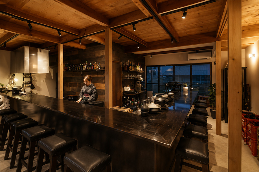 A long dark wooden bar with black cushioned stools, bartender at work, shelves of bottles behind, warm overhead lighting, and a windowed back wall.