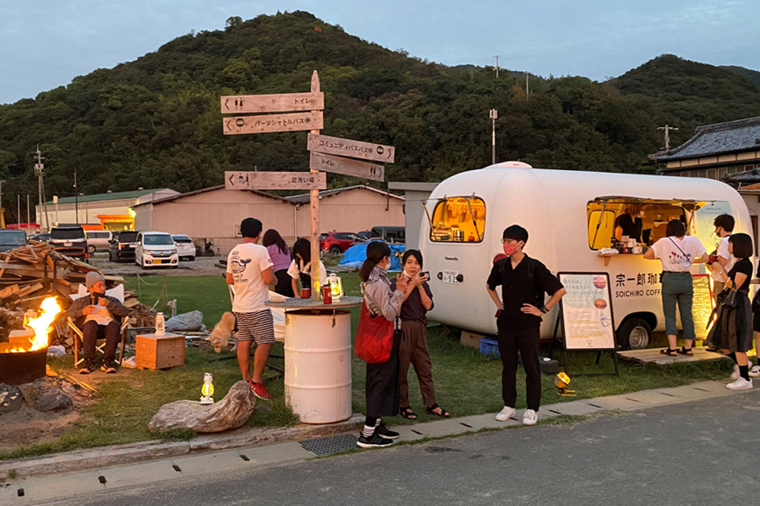 Group of people gathered around a campfire near a white food-truck van at dusk, with a wooden signpost and outdoor seating area behind them.