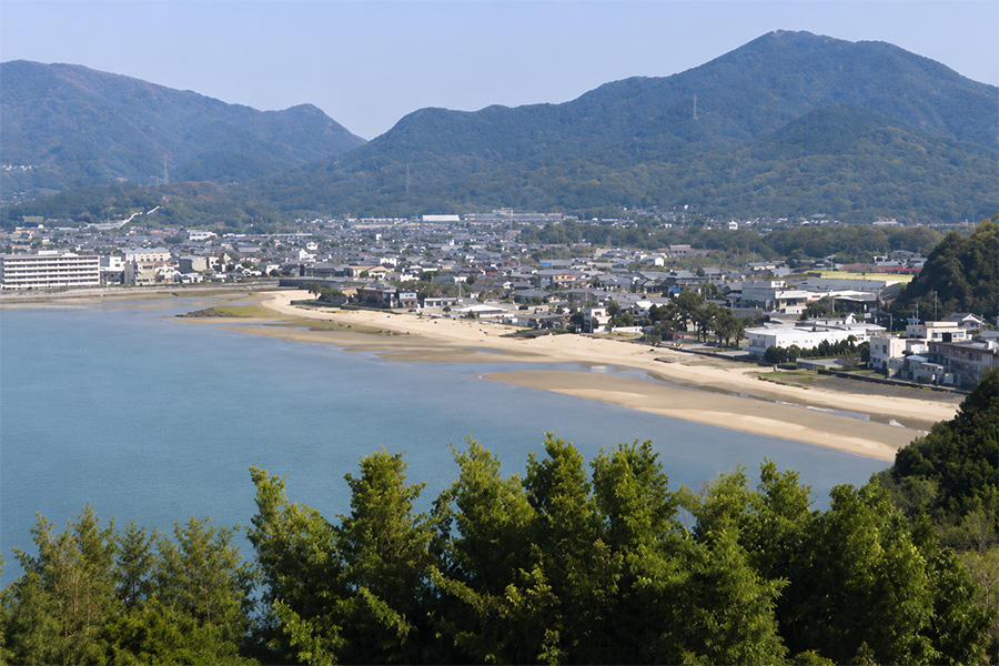 A seaside town along a curved sandy bay with calm blue water and green hills in the background.