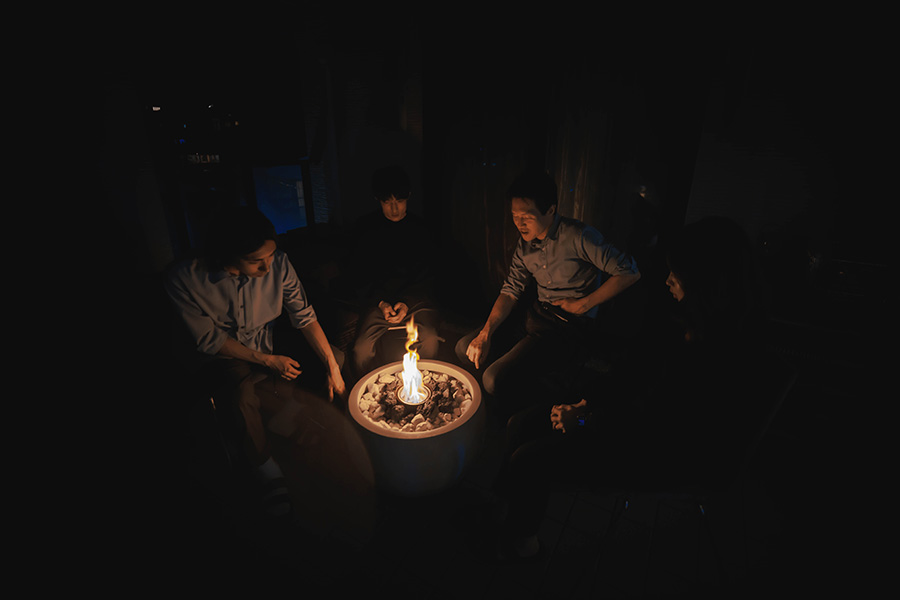 Five people sit around a lit fire pit in a dark setting, the flames casting a warm glow on their faces.