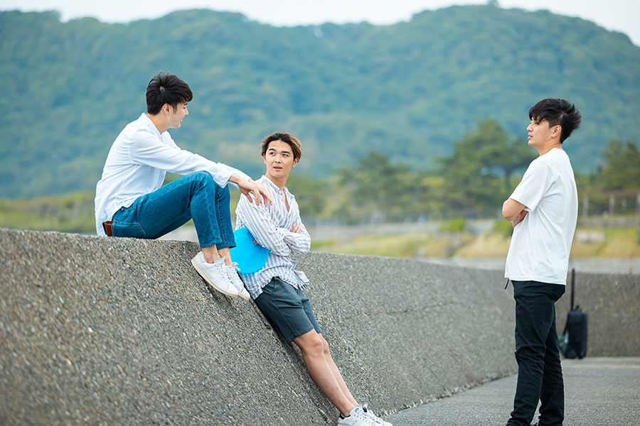 Three young men by a concrete wall near a scenic hillside; two sit and chat while the third stands nearby, listening.