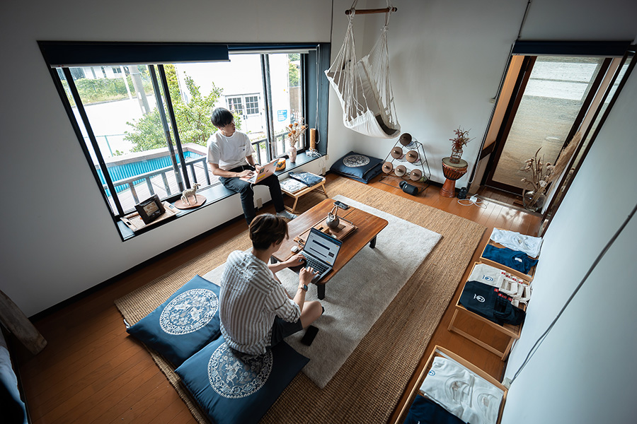 Two people work on laptops in a sunlit loft living room with a low wooden table, floor cushions, and a hanging hammock chair.