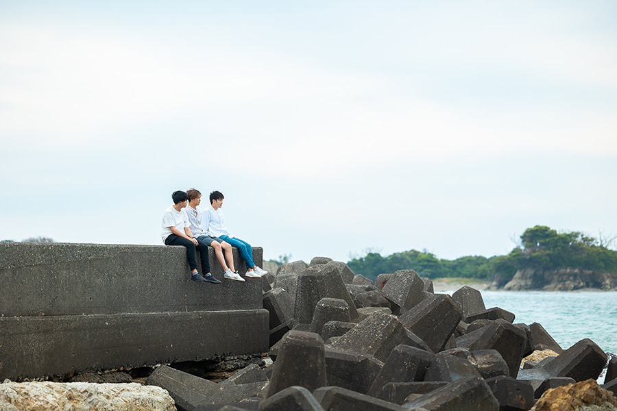Four young friends sit on a concrete seawall overlooking the water, with rocky breakwater stones in the foreground and a distant shoreline.