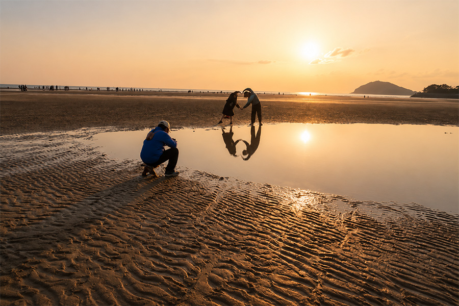 Couple forms a heart with their arms over a reflective pool at sunset on a sandy beach, photographer crouched nearby.