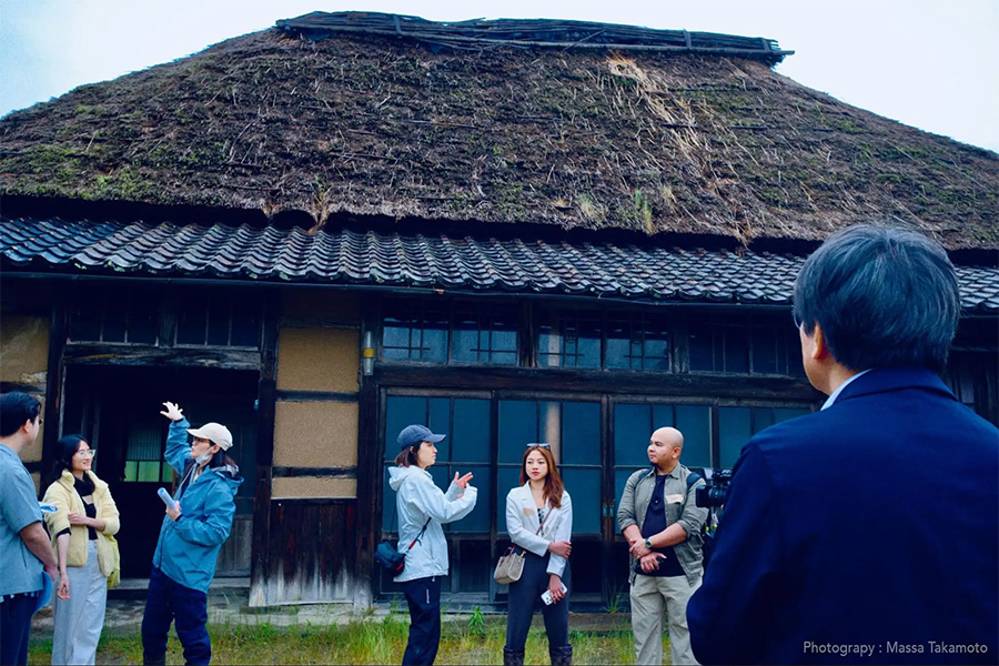 People gather outside a rustic thatched-roof building as a video crew films a tour or interview.