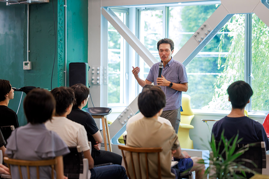 Man speaks into a microphone to a seated audience in a bright, industrial-style room with large windows.