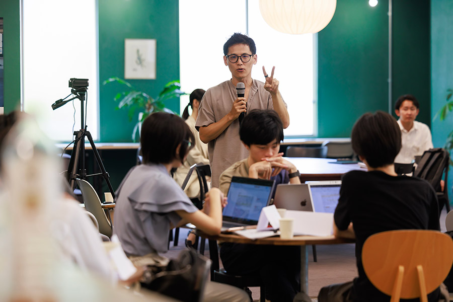 Man with glasses speaks into a microphone at a round-table workshop, gesturing a peace sign to participants in a bright room: informative event scene.