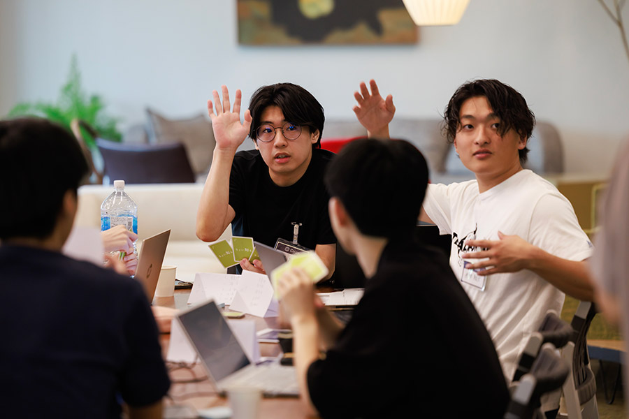 A group of people around a table during a workshop, two men raise their hands to answer a question.