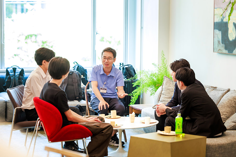 A group of five coworkers sitting in a bright lounge area, chatting around a coffee table with cups and snacks.