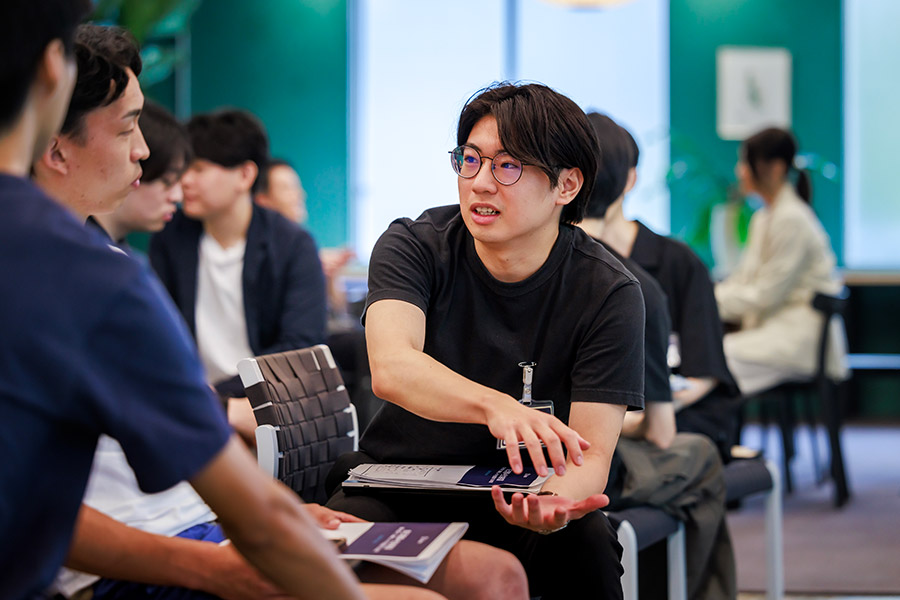 Young man with glasses explains something to a group during a workshop, gesturing with his hand while seated among teammates.