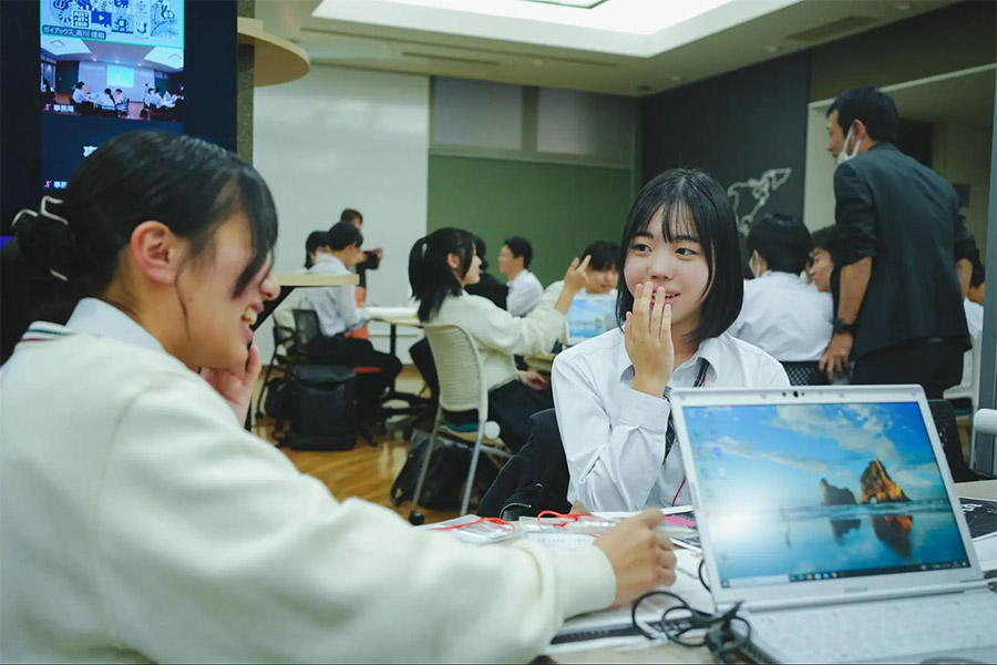 Two students sit at a table in a classroom with laptops, one smiling and the other touching her face.