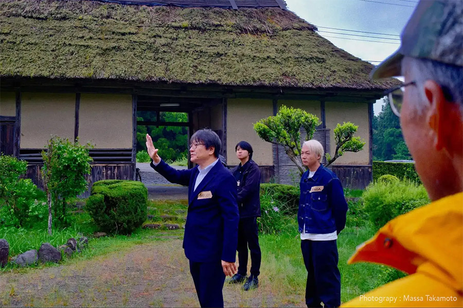 Man in a dark blue suit raises his hand while leading a small group in a garden in front of a thatched-roof house.