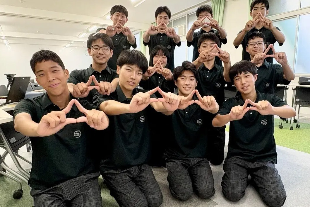 Twelve students in black shirts pose in a classroom, forming triangle shapes with their fingers and smiling at the camera.